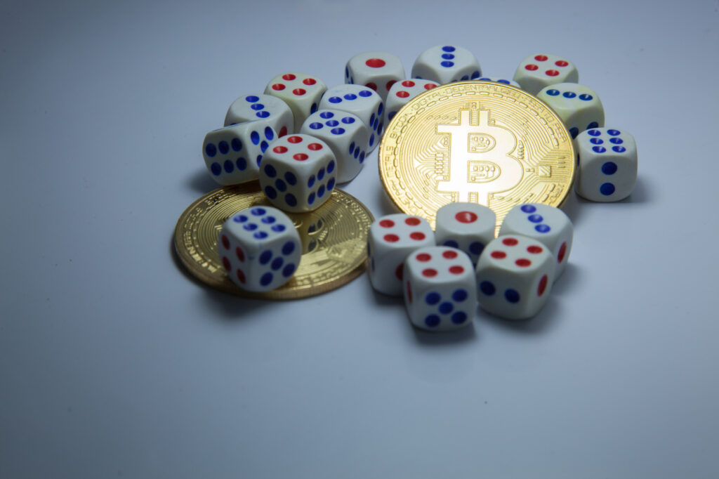 A gold Bitcoin coin, symbolizing the allure of VPN-friendly Bitcoin casinos, is surrounded by several white dice adorned with red and blue dots on a light surface. | FintechZoom