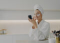 A woman in a white robe and towel on her head is smiling at her phone, perhaps reading skin care tips. She has eye patches under her eyes and is sitting in a modern bathroom, with beauty products visible in the foreground. | FintechZoom