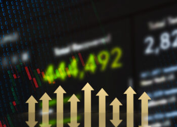 Blurred financial data and stock charts with green numbers indicating upward movement. A series of gold upward arrows are prominently displayed in the foreground, symbolizing growth or increase in value. | FintechZoom