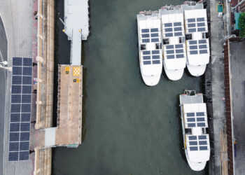An aerial view captures five boats docked in the harbor, each outfitted with sleek solar panels on their roofs. A pier, also adorned with solar panels, extends into the water from a paved walkway lined with streetlights, showcasing a commitment to renewable energy. | FintechZoom