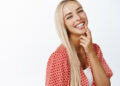 A woman with long blonde hair smiles joyfully, showcasing a radiant smile that hints at the confidence from a recent dental implant. She wears a red and white patterned blouse over a white top, touching her chin with one finger against a plain white background. | FintechZoom
