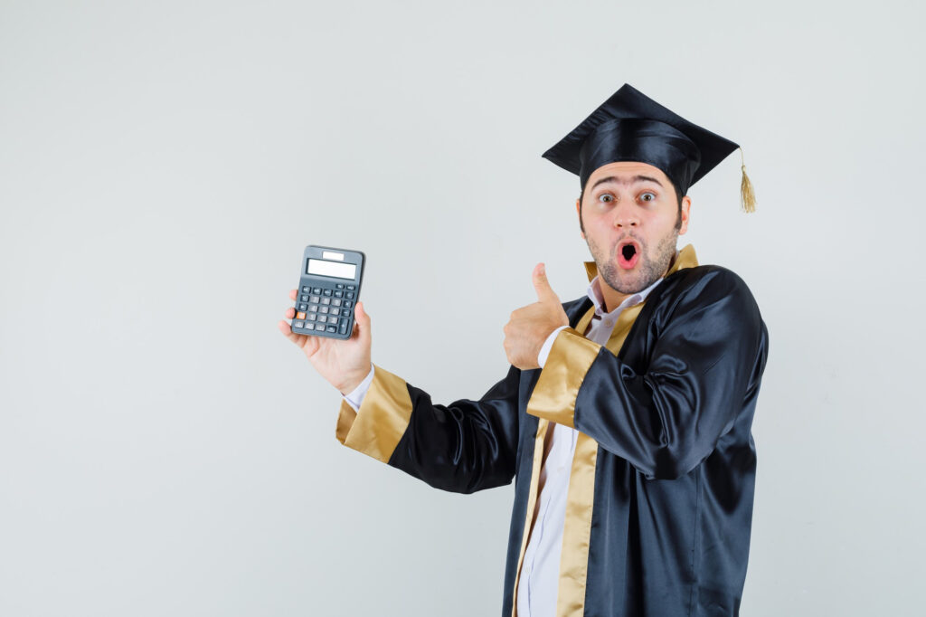 A young man in a graduation cap and gown clutches a student loan calculator in one hand, giving a thumbs-up with the other, his face radiating surprise and excitement. The plain light gray background accentuates his achievement. | FintechZoom