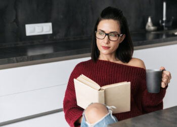 A woman wearing glasses and a red sweater sits in a modern kitchen, deeply engrossed in a book. With a gray mug in hand, she appears relaxed yet focused, as if preparing for the next challenge of the Cerebrum IQ test. Her dark, straight hair frames her contemplative expression perfectly. | FintechZoom