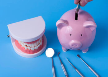 A piggy bank with a coin being inserted is placed next to a set of model teeth. Below them are four dental tools: a mirror, a probe, and two picks, all arranged on a blue background. | FintechZoom