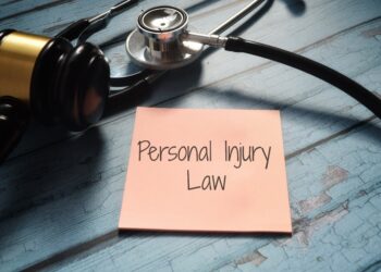 A stethoscope and a gavel are placed on a blue wooden surface next to a pink sticky note with the words "Personal Injury Law" written on it. | FintechZoom