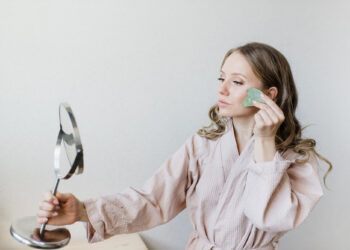 A woman in a light pink robe embraces her beauty routine, using a jade roller on her face while gazing into a handheld mirror. Her long, wavy hair complements the minimalist setting surrounding her. | FintechZoom