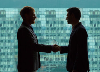 Two people in business attire are shaking hands, symbolizing strong business relationships, in front of a large window filled with square panes. Their silhouetted figures create a striking contrast against the light streaming through the glass. | FintechZoom
