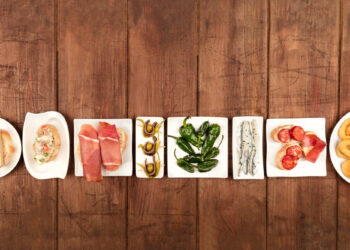 A variety of tapas on white plates is arranged in a line on a wooden surface, tempting even those practicing fasting. The dishes feature meats, peppers, anchovies, tomatoes, and pastries. | FintechZoom