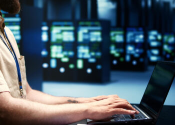 In a dimly lit room, a person types on a laptop, surrounded by numerous server racks hinting at the perfect data center environment. Wearing a light-colored shirt and blue lanyard, they navigate the best VPS for futures trading with focused precision. | FintechZoom