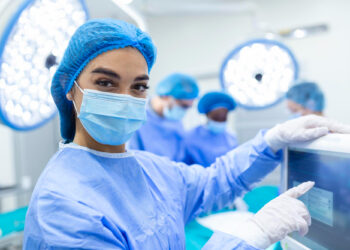 A medical professional in surgical attire, including a blue gown, hair cap, mask, and gloves, stands near a screen in an operating room. Three others in similar attire are in the background, working together. | FintechZoom