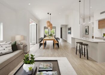 Modern open-plan living space with a kitchen, dining area, and living room. The room features light wood flooring, a large dining table with chairs, pendant lights, a gray sofa, a coffee table, and ample natural light from a window and glass door. | FintechZoom