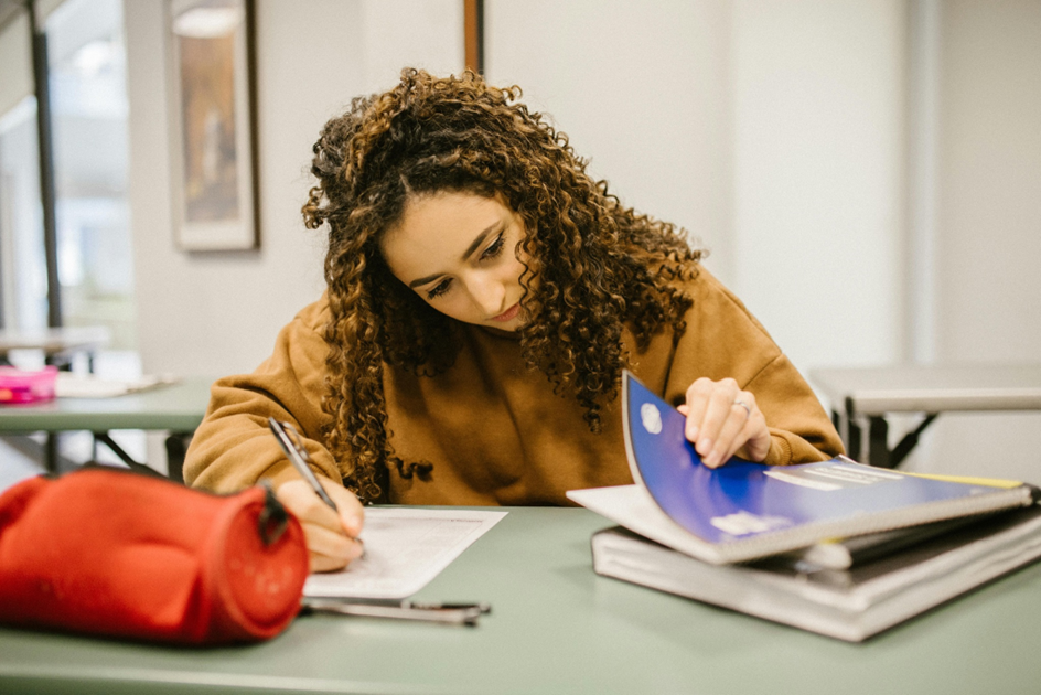 A woman with curly hair is sitting at a table, writing in a notebook. She is wearing a brown sweater and is focused on her work. There is a red pencil case and other books on the table. The setting appears to be a classroom or library. | FintechZoom