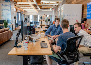 Several people are engaged in a discussion at a modern office. Three men are seated at a desk with computers and other office equipment around. A few others are working or walking in the background. The atmosphere appears collaborative. | FintechZoom