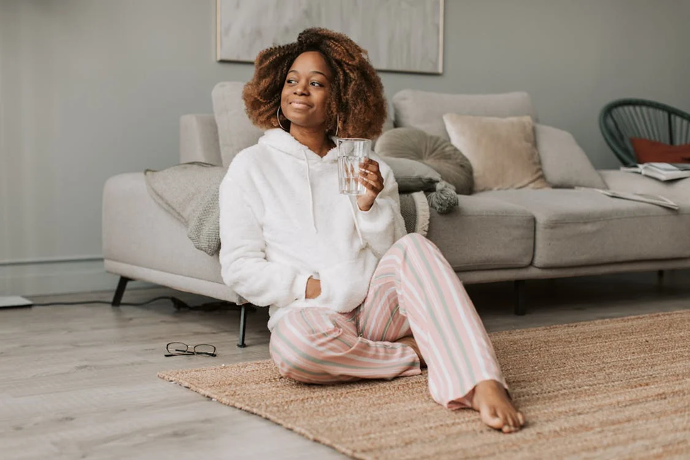 A person sitting on a rug in a cozy living room, wearing a white fluffy hoodie and striped pajama pants. They are holding a glass of water and smiling. The background features a sofa with cushions and a wall painting. | FintechZoom