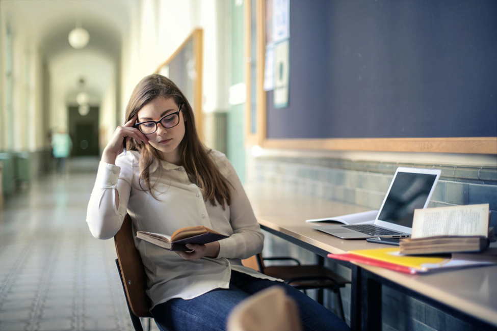 A woman with glasses sits at a table in a hallway, reading a book. An open laptop, a stack of books, and notebooks are on the table. The walls are lined with notice boards, and the corridor stretches into the distance. | FintechZoom