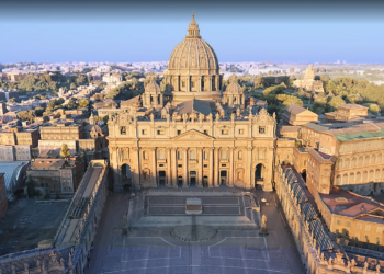 Aerial view of St. Peter's Basilica in Vatican City, showcasing its iconic dome and grand facade. The surrounding area includes historic buildings and greenery under a clear blue sky, capturing the heart of the Vatican. | FintechZoom