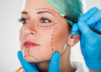 A cosmetic surgeon wearing a surgical cap and blue gloves marks dotted lines on a woman's face with a pencil, highlighting areas for potential cosmetic treatment. The woman gazes forward with a neutral expression. | FintechZoom