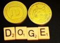 Two gold Dogecoin tokens symbolize the rise of meme coins; one displays the iconic "D" logo, while the other features a Shiba Inu and "WOW". Below them, Scrabble tiles spell "DOGE" against a sleek black background. | FintechZoom