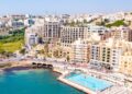Aerial view of Malta city with a prominent shoreline swimming pool labeled "Surfside Lido." This picturesque hub for residential property investment in Malta boasts multi-story buildings and a road curving along the waterfront, while greenery adorns structures under a clear blue sky. | FintechZoom
