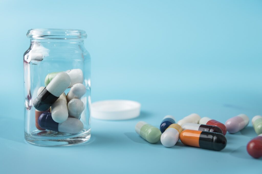 A clear glass jar filled with various colorful capsules is open, with more capsules scattered on a light blue surface. A white jar lid is placed nearby, creating a vibrant and medical-themed composition. | FintechZoom