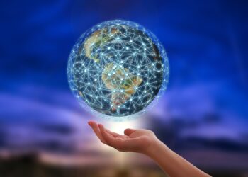 A hand is extended upward, appearing to support a glowing digital globe, embodying the essence of blockchain technology. The globe is overlaid with interconnected lines and dots against a blurred, dark blue and purple sky, evoking a sense of evening or night. | FintechZoom