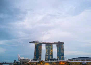 The image shows Marina Bay Sands in Singapore, with its three tall towers supporting a ship-like structure on top. The sky is partly cloudy, and there are other buildings nearby, including a lotus-shaped structure. | FintechZoom