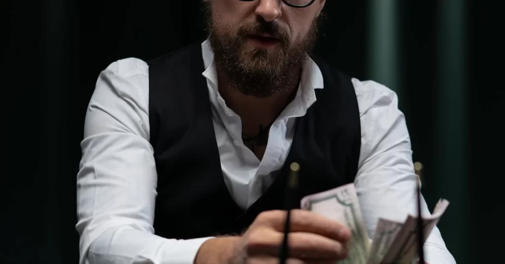 A bearded man, wearing glasses and a white shirt with a black vest, sits with a serious demeanor, counting a stack of dollar bills. The dark background amplifies the intensity, hinting at his recent success in offshore casino games. | FintechZoom