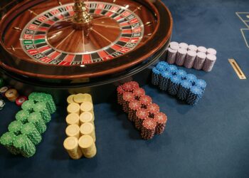 A roulette wheel on a casino table showcases stacks of poker chips in green, yellow, red, blue, and purple neatly arranged beside it—an enticing setup for testing your casino strategies. The table is elegantly draped in blue felt fabric. | FintechZoom