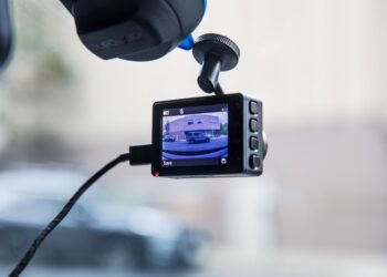 A dash board camera mounted on a windshield records the road ahead. The screen shows a car in its view. The device is connected by a visible cable. The background is blurred, indicating motion or focus on the camera. | FintechZoom