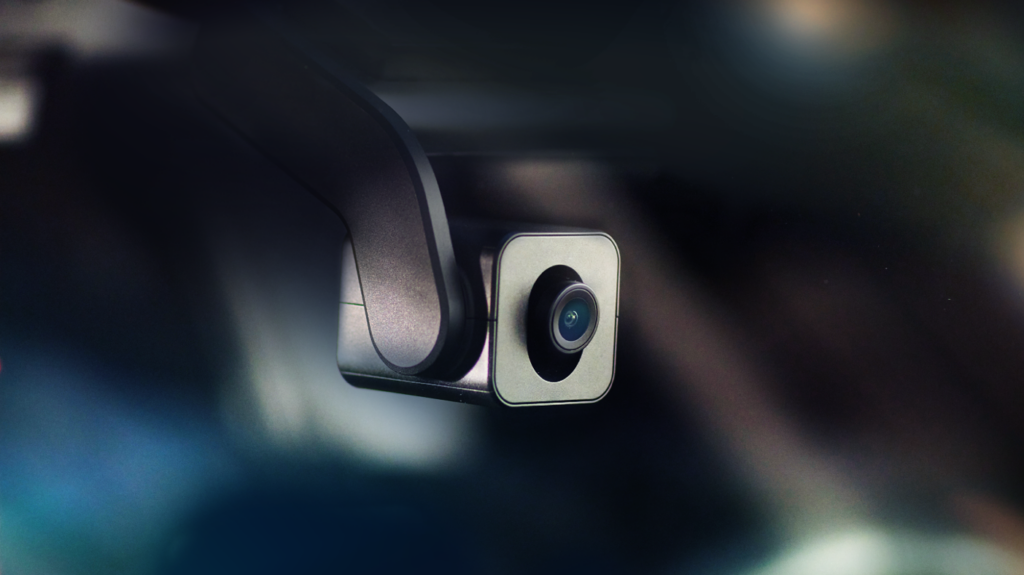 Close-up of a compact, modern dashcam mounted inside a vehicle, with a sleek design and dark metallic finish. The camera lens is prominently visible, and the background is blurred, emphasizing the device. | FintechZoom