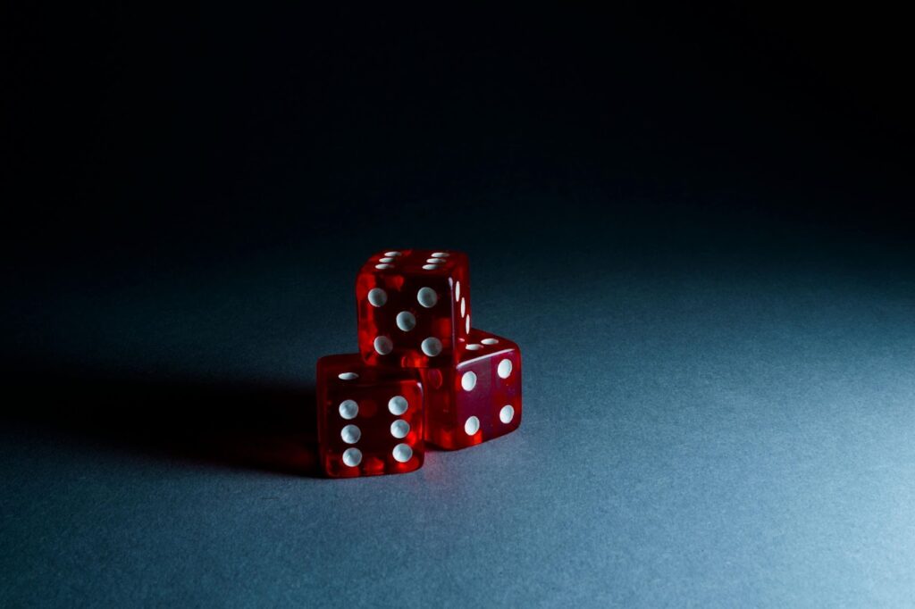 Casino innovations encourage gambling ETF - Three red dice with white dots are stacked on a gray gradient surface. The lighting highlights the translucent quality of the dice, casting soft shadows underneath. | FintechZoom