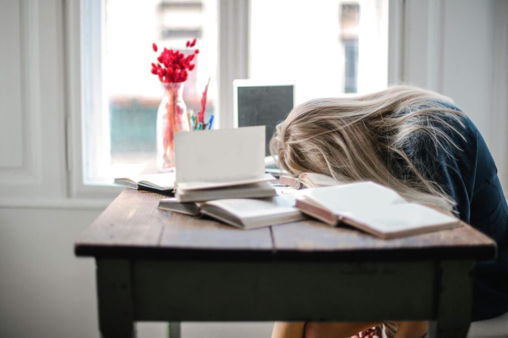 A person with long hair sits at a wooden desk with their head resting on an open book. Several other books and papers are scattered on the desk. A vase with red flowers is in the background near a window. | FintechZoom