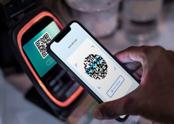 A person is holding a smartphone displaying a QR code on its screen, positioned next to a payment terminal. The terminal's display also shows a QR code, highlighting the modern evolution of payment methods through seamless contactless transactions. | FintechZoom