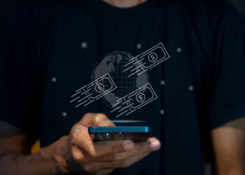A person holds a smartphone, with a digital illustration of a globe and dollar bills floating above, symbolizing global money transfer. The scene is reminiscent of how FintechZoom.com tracks the rise of neobanks and digital wallets. The individual dons a dark shirt. | FintechZoom