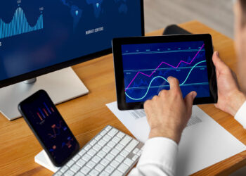 A person at a desk intensely analyzes financial charts on a tablet, while FintechZoom.com reports flash across the computer monitor. Nearby, a smartphone displays more charts amid the clutter of a keyboard and scattered papers on the wooden surface. | FintechZoom
