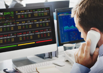 A person talks on a phone at a desk, with computer monitors displaying data from Fintechzoom.com. The screens show different global stock indices and financial metrics in various colors, indicating market trends. | FintechZoom