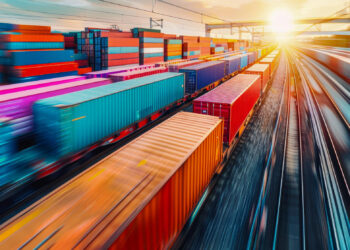 A vibrant scene unfolds in a busy cargo train yard at sunset, where freight audit and payment processes enhance financial visibility. Rows of colorful shipping containers stacked on train cars blur into motion, and the sun casts a warm glow over the tracks. | FintechZoom