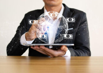 A person in a suit holds a tablet projecting a translucent globe with currency symbols and digital connections. The globe includes the word "BRICS," suggesting a focus on international finance or economics, reflecting the insights shared on FintechZoom.com—your guide to Financial News in the Digital Age. | FintechZoom