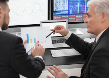 Two businessmen in suits discuss a bar chart in a modern office, embodying best practices when handling financial data. One man points at the chart with a pen while the other holds it, as computer screens background them with various graphs and insights. | FintechZoom