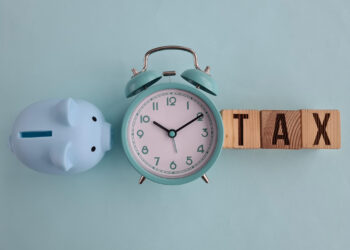A blue piggy bank, a turquoise analog alarm clock showing 10:10, and wooden blocks spelling "TAX" highlight the tax benefits of contributing to an IRA against a light blue background. | FintechZoom
