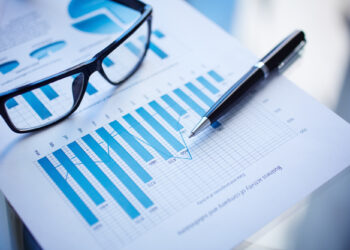 A pair of eyeglasses and a pen rest on a sheet of financial graphs, alongside a balance sheet template. The charts feature blue bar graphs and line plots, showcasing numerical data and trends. The scene suggests an analysis or review of business or financial reports. | FintechZoom