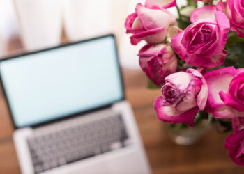 A blurred open laptop on a wooden surface with a bright white screen is in the background, and a clear close-up of a bouquet of vibrant pink roses is in the foreground on the right side. | FintechZoom