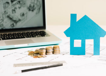 A paper cutout of a blue house stands on a table cluttered with architectural plans, coins, and a ruler—symbols of dreams funded by home improvement loans. A laptop displaying a blueprint is partially visible in the background. | FintechZoom