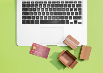 A laptop on a green background with a red credit card and two small open cardboard boxes nearby. The image suggests online shopping or e-commerce. | FintechZoom