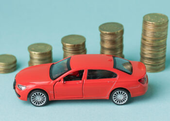 A red toy car sits on a blue background, flanked by four stacks of coins that grow taller from left to right, signifying financial growth or car expenses like car loans. | FintechZoom