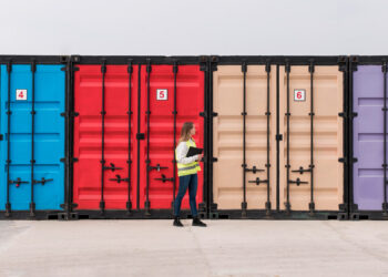 A person in a yellow safety vest stands before a row of colorful, refrigerated shipping containers, clipboard in hand. Painted blue, red, beige, and purple, these containers are integral to logistics and marked with numbers 4, 5, and 6. | FintechZoom