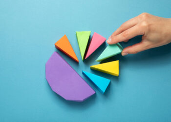 A hand arranges colorful triangular pieces of paper into a circular design on a blue surface, much like the intricate balance of tokenomics. The pieces are orange, green, pink, light green, yellow, and blue, with a larger purple piece completing the formation. | FintechZoom