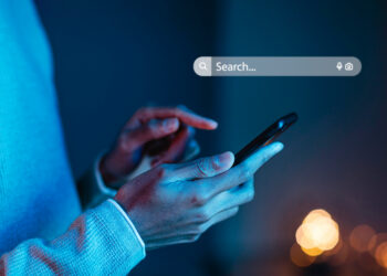 A person in a light sweater types on a smartphone, framed by the warm glow of blurred background lights. A search bar floats above, symbolizing internet exploration—perhaps just as Google announced a new rule changing the world of online data processing. | FintechZoom