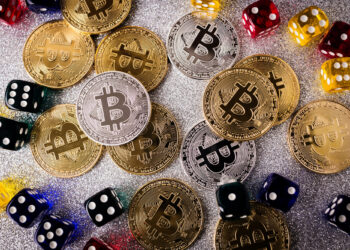 A scattered arrangement of gold and silver Bitcoin coins on a sparkly surface, surrounded by colorful dice in red, yellow, green, blue, and black invites you to imagine the thrill of betting with cryptocurrencies. The coins display the Bitcoin logo prominently. | FintechZoom