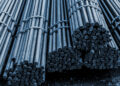 Bundles of steel rebar are stacked together, with the ends facing outward. The rods are tied with metal straps, displaying a textured, cylindrical surface. The image is captured with a blue tint, highlighting the industrial setting. | FintechZoom